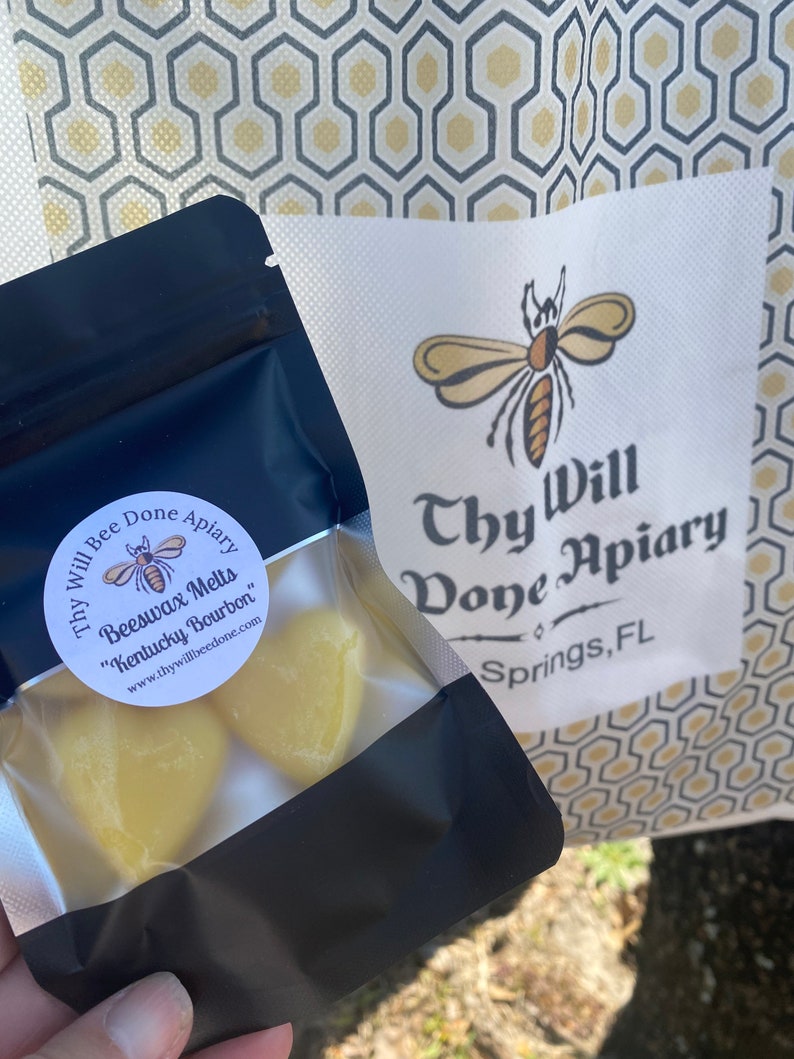 Może przedstawiać: Czarna zamykana torebka zawierająca ż&oacute;łte woski pszczele w kształcie serca z etykietą Thy Will Bee Done Apiary Kentucky Bourbon. Obok znajduje się biała papierowa torba z logo pszczoły i napisem Thy Will Bee Done Apiary Springs, FL.