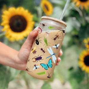 May include: A glass tumbler with a bamboo lid and a clear straw, featuring a beige background adorned with colorful insect illustrations. The tumbler is filled with a light-colored beverage.