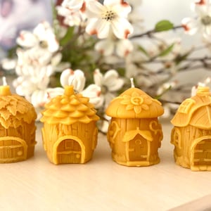 Peut inclure: Quatre petites bougies en cire d'abeille en forme de maisons, aux détails complexes. Chaque maison a un design différent, y compris un toit en forme de champignon, un toit en forme de pomme de pin et un toit en chaume. Les bougies sont de couleur jaune clair et sont disposées en rangée sur une surface en bois.