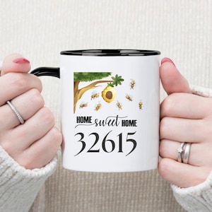 May include: White ceramic mug with a black rim and handle. The mug features a cartoon illustration of a beehive with bees flying around it. The text "HOME sweet HOME" and the number "32615" are printed on the mug.