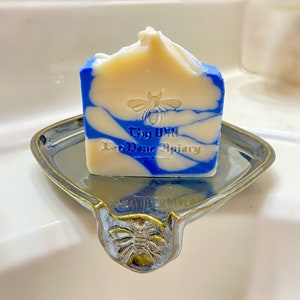 May include: A blue and white swirl soap bar with a bee and the text "Thy Will Bee Done Apiary" on a ceramic soap dish with a bee design.