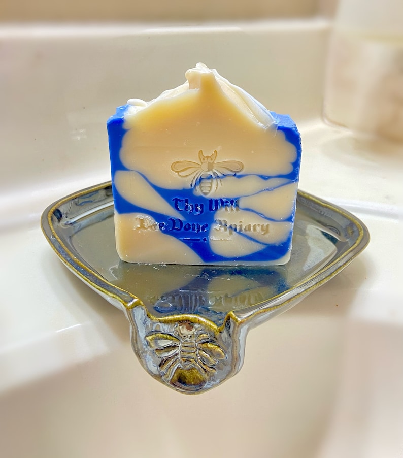 May include: A blue and white swirl soap bar with a bee and the text "Thy Will Bee Done Apiary" on a ceramic soap dish with a bee design.