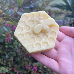 May include: A pale yellow hexagonal bar of soap featuring a raised relief design of a honeybee centred on a honeycomb pattern. The soap has a smooth, matte finish and is shaped like a single honeycomb cell cluster with defined geometric edges.