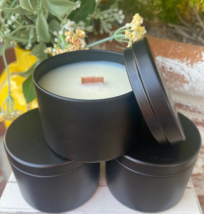 Pu&ograve; includere: Un set di tre barattoli per candele in metallo nero opaco, con uno aperto che rivela cera bianca e uno stoppino piatto in legno. Il coperchio del barattolo aperto &egrave; appoggiato di lato. I contenitori sono cilindrici con superfici lisce.