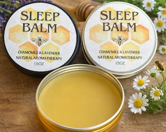 Op de afbeelding: Twee ronde metalen blikjes slaapbalsem met zwarte en zilveren deksels, elk voorzien van een honingraatpatroon, een bijenillustratie en de tekst Sleep Balm Chamomile & Lavender Natural Aromatherapy 1.5oz. Een open blikje toont gele balsem.