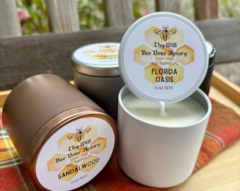 May include: Three 3 oz soy candles in metal tins. One copper-coloured tin is labelled Sandalwood, while a white tin features a lid labelled Florida Oasis with a bee and honeycomb graphic. A third tin in the back is labelled Cranberry Honey.