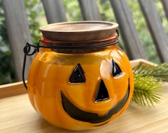 Puede incluir: Un tarro de cristal naranja con forma de calabaza de Halloween con una cara pintada en negro, ojos triangulares y una boca ancha sonriente. El tarro tiene un asa de alambre negro fino y una tapa circular de madera en la parte superior.