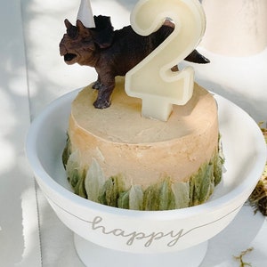 May include: A small birthday cake with a brown triceratops figurine wearing a party hat and a number 2 candle. The cake sits in a white pedestal bowl with the word "happy" written on the side.