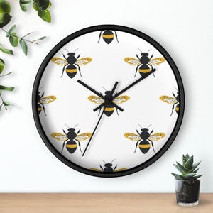 May include: A round wall clock with a black frame and a white face. The clock face features a repeating pattern of stylized yellow and black bees. The clock hands are black. The clock is on a wooden surface.