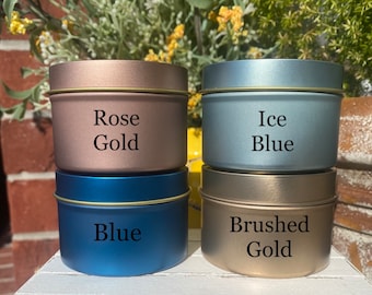 May include: Four cylindrical metal tins stacked in two pairs. The top left tin is rose gold, the top right is ice blue, the bottom left is blue, and the bottom right is brushed gold. Each tin features its respective colour name printed in black text.