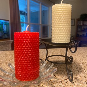 May include: A red cylindrical honeycomb-textured candle sits in a clear glass flower-shaped dish, while a cream-colored cylindrical honeycomb-textured candle rests on a black metal stand with curved legs and a circular base.
