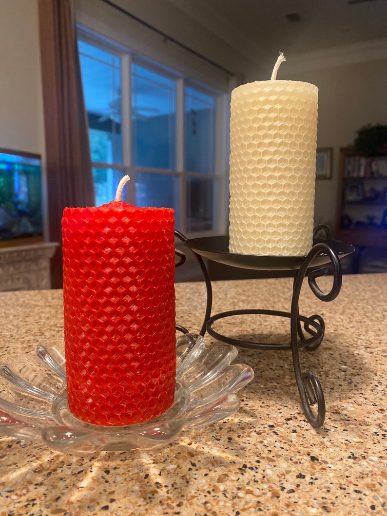 May include: A red cylindrical honeycomb-textured candle sits in a clear glass flower-shaped dish, while a cream-colored cylindrical honeycomb-textured candle rests on a black metal stand with curved legs and a circular base.