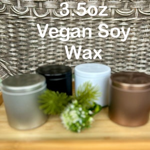 May include: Four cylindrical metal tins containing 3.5oz Vegan Soy Wax, featuring silver, black, white, and bronze finishes with matching lids. A small green decorative plant sprig is placed in the center among the containers.