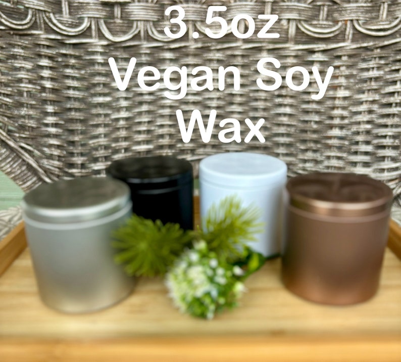 May include: Four cylindrical metal tins containing 3.5oz Vegan Soy Wax, featuring silver, black, white, and bronze finishes with matching lids. A small green decorative plant sprig is placed in the center among the containers.