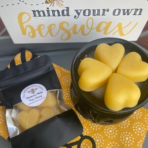 Może przedstawiać: Czarny podgrzewacz do wosku z czterema ż&oacute;łtymi woskami pszczelimi w kształcie serca, obok czarnej torebki z etykietą Thy Will Bee Done Apiary Beeswax Melts Pure Honey oraz białego znaku z ilustracją pszczoły i napisem mind your own beeswax.