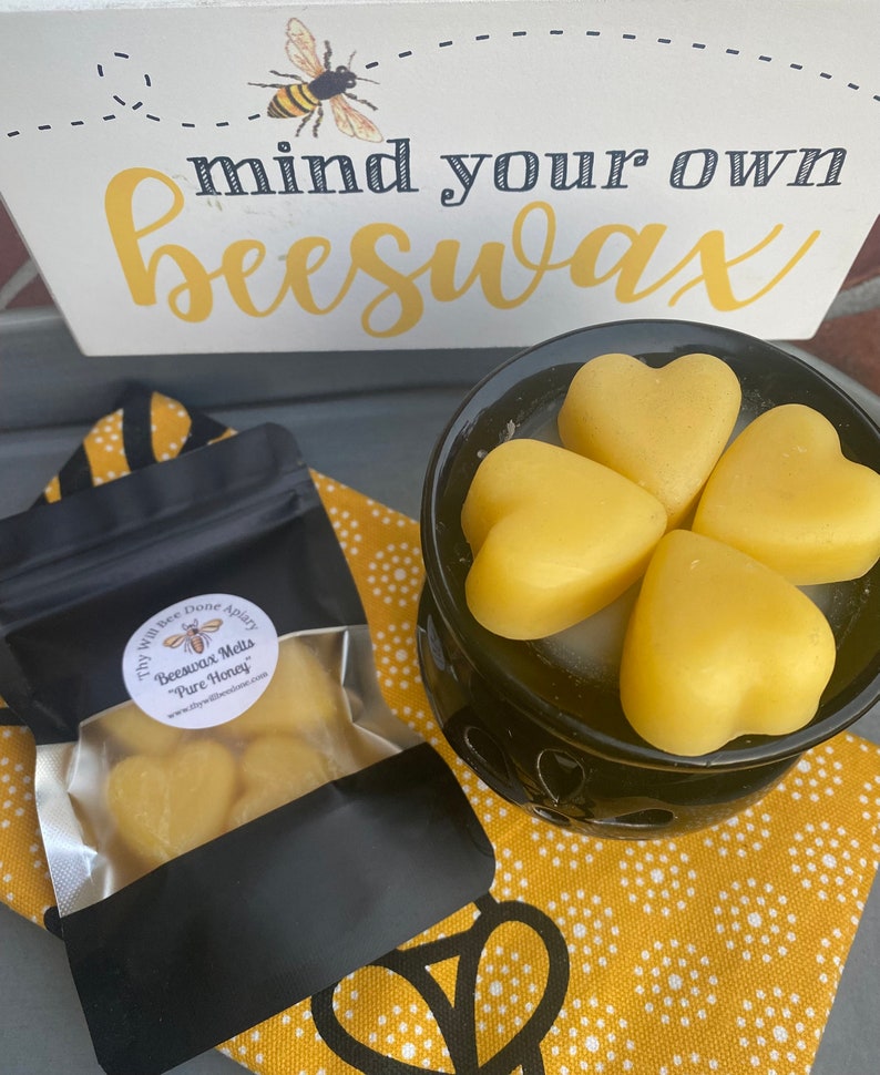 Może przedstawiać: Czarny podgrzewacz do wosku z czterema ż&oacute;łtymi woskami pszczelimi w kształcie serca, obok czarnej torebki z etykietą Thy Will Bee Done Apiary Beeswax Melts Pure Honey oraz białego znaku z ilustracją pszczoły i napisem mind your own beeswax.