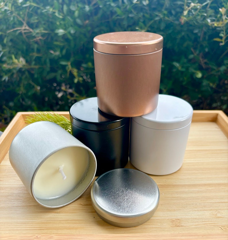 May include: A collection of four cylindrical metal candle tins in rose gold, matte black, white, and silver. One silver tin is open, revealing a white wax candle with a centered wick, while its matching silver lid rests in front of the group.