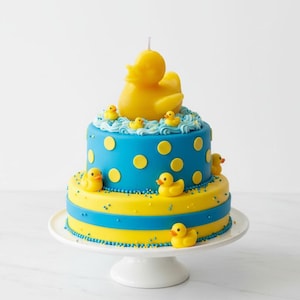 May include: A two-tier cake decorated with blue and yellow fondant, featuring yellow polka dots, blue frosting swirls, and multiple small yellow rubber duck figures. A large yellow rubber duck candle sits on the top tier of the cake.
