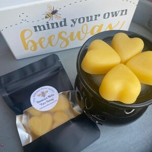 Może przedstawiać: Czarny podgrzewacz do wosku z czterema ż&oacute;łtymi woskami pszczelimi w kształcie serca, czarna torebka z etykietą Thy Will Bee Done Apiary Beeswax Melts Pure Honey oraz biały znak z grafiką pszczoły i napisem mind your own beeswax.
