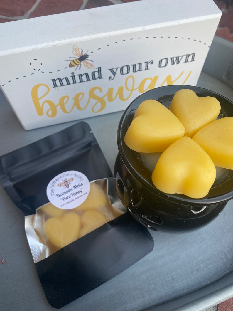Może przedstawiać: Czarny podgrzewacz do wosku z czterema ż&oacute;łtymi woskami pszczelimi w kształcie serca, czarna torebka z etykietą Thy Will Bee Done Apiary Beeswax Melts Pure Honey oraz biały znak z grafiką pszczoły i napisem mind your own beeswax.