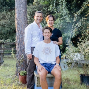 May include: A family of three poses outdoors, with a young person seated on a blue and white box. The person wears a white t-shirt with a circular graphic, blue shorts, and white sneakers. The parents stand behind, near a tree.