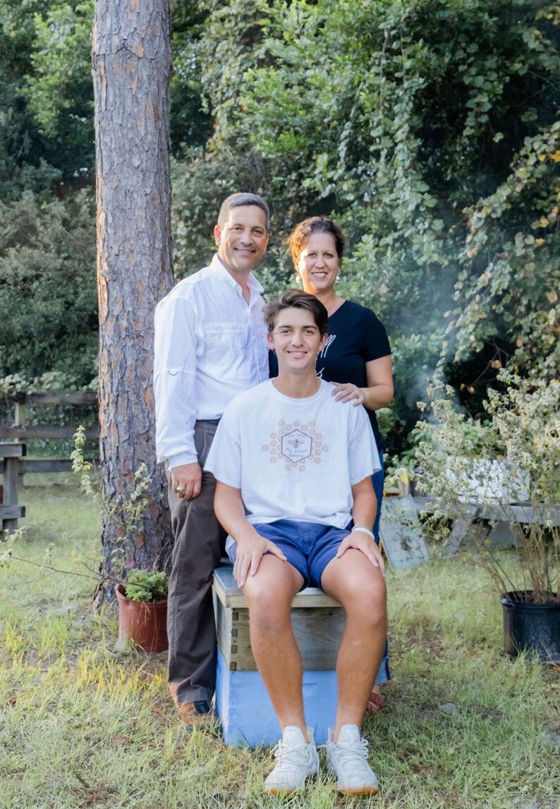 May include: A family of three poses outdoors, with a young person seated on a blue and white box. The person wears a white t-shirt with a circular graphic, blue shorts, and white sneakers. The parents stand behind, near a tree.