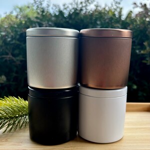 May include: Four cylindrical metal tins with lids arranged in a two-by-two stack. The top left tin is silver, the top right is bronze, the bottom left is matte black, and the bottom right is white. Each container has a smooth, solid finish.