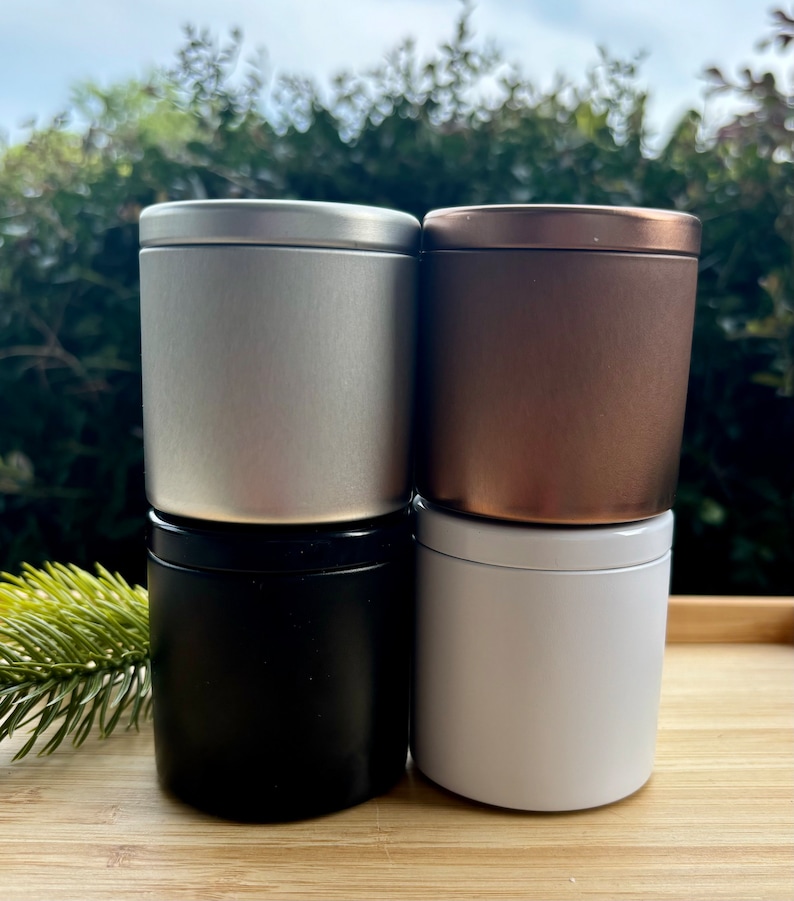 May include: Four cylindrical metal tins with lids arranged in a two-by-two stack. The top left tin is silver, the top right is bronze, the bottom left is matte black, and the bottom right is white. Each container has a smooth, solid finish.