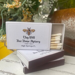 May include: White matchboxes with brown edges are stacked on a white wooden surface. The top box is open, revealing several matches with white tips. The boxes feature a bee logo and the text "Thy Will Bee Done Apiary, High Springs, FL."