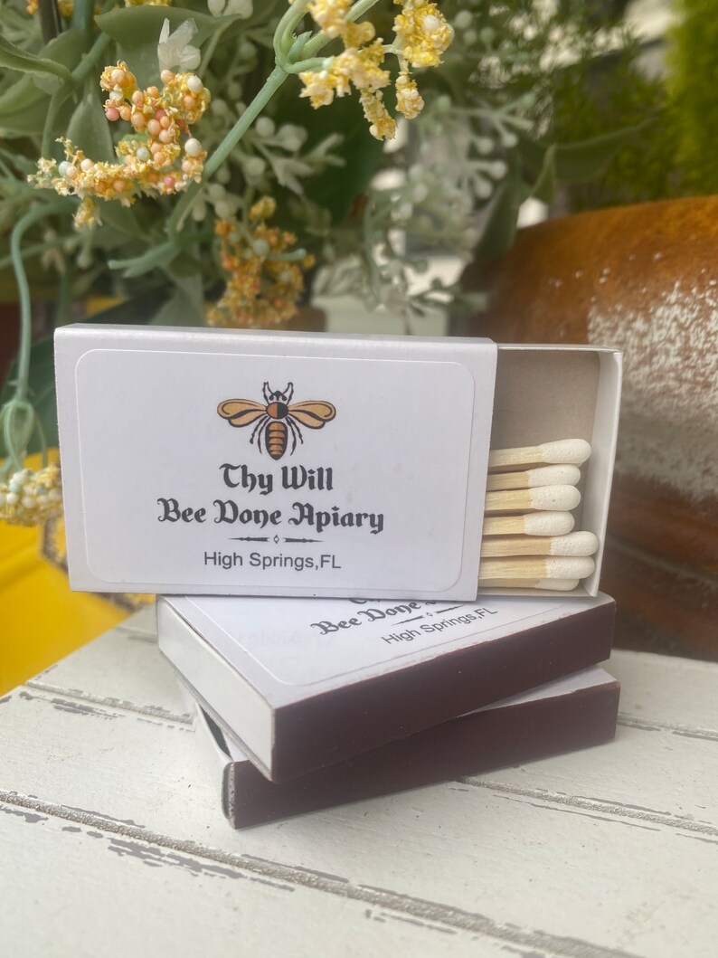 May include: White matchboxes with brown edges are stacked on a white wooden surface. The top box is open, revealing several matches with white tips. The boxes feature a bee logo and the text "Thy Will Bee Done Apiary, High Springs, FL."
