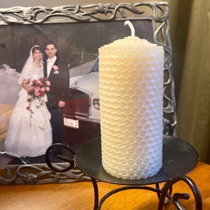 May include: A cylindrical off-white candle with a textured honeycomb pattern on its surface. The candle features a single cotton wick at the top and is positioned upright on a black metal candle holder with curved legs and a circular base.