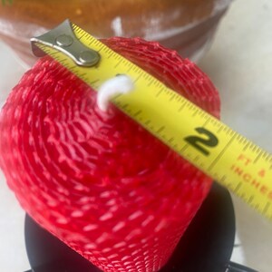 May include: A red cylindrical beeswax candle with a textured honeycomb pattern and a white wick, positioned in a black metal candle holder. A yellow measuring tape is placed across the top, showing a diameter of approximately 2 inches.