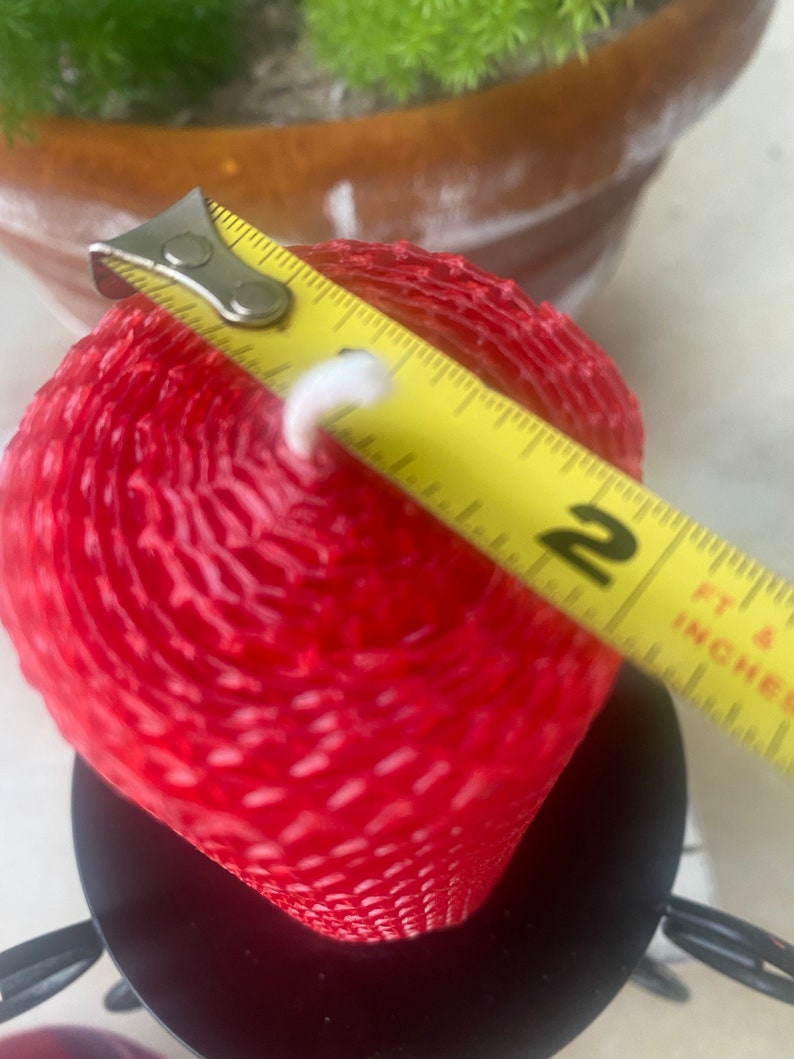 May include: A red cylindrical beeswax candle with a textured honeycomb pattern and a white wick, positioned in a black metal candle holder. A yellow measuring tape is placed across the top, showing a diameter of approximately 2 inches.