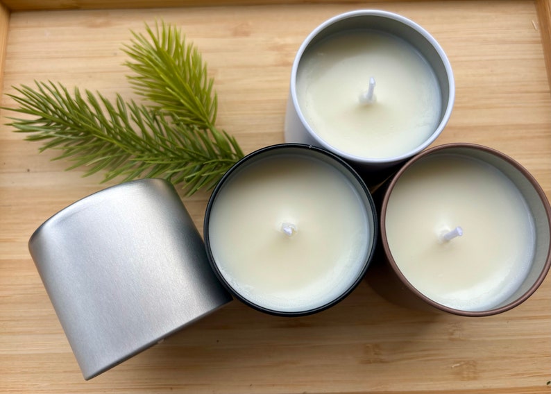 May include: Three round metal candle tins containing white wax with centered wicks. One tin is silver, one is black, and one is bronze. A small green artificial pine sprig rests beside the silver tin, which is shown from the side.