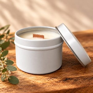 May include: A round, light gray metal tin containing a white candle with a rectangular wooden wick. The matching circular metal lid is resting on its side next to the container. The candle wax is smooth and fills the tin to the top edge.