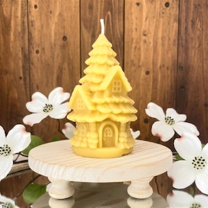 May include: A yellow candle shaped like a house nestled within a pine tree, featuring a central arched doorway, small windows, and a textured roof. The candle has a single white wick at the top and sits on a round, light-colored wooden pedestal.