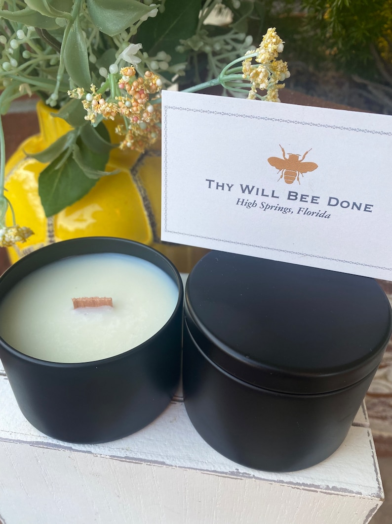 Pu&ograve; includere: Due barattoli per candele in metallo nero opaco, uno aperto che rivela cera bianca con uno stoppino in legno e uno chiuso con un coperchio. Un biglietto da visita rettangolare bianco presenta un'icona di ape dorata e il testo Thy Will Bee Done High Springs, Florida.