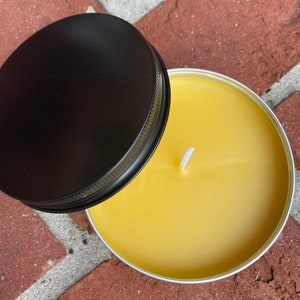May include: A round, yellow candle in a silver tin with a black lid. The candle has a white wick and is set against a brick background. The lid is partially open, revealing the candle's surface.