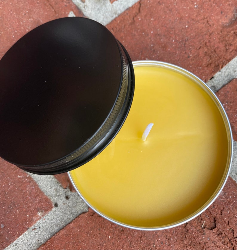 May include: A round, yellow candle in a silver tin with a black lid. The candle has a white wick and is set against a brick background. The lid is partially open, revealing the candle's surface.