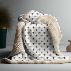 May include: A white blanket with a repeating pattern of gold and black bees. The blanket has a soft, cream-colored sherpa lining. The blanket is folded over a surface, showcasing the bee design and the plush texture.