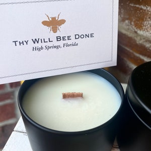 Pu&ograve; includere: Una candela bianca con stoppino in legno in un barattolo di metallo nero opaco. Un cartoncino bianco rettangolare presenta un'icona di ape dorata e il testo "THY WILL BEE DONE" seguito da "High Springs, Florida" con un bordo decorativo.