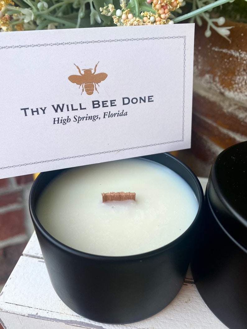 Pu&ograve; includere: Una candela bianca con stoppino in legno in un barattolo di metallo nero opaco. Un cartoncino bianco rettangolare presenta un'icona di ape dorata e il testo "THY WILL BEE DONE" seguito da "High Springs, Florida" con un bordo decorativo.