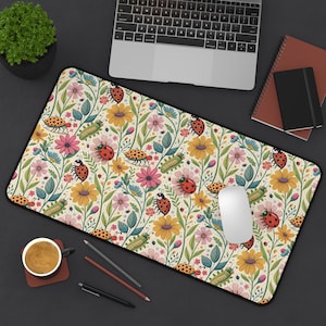 May include: A rectangular desk mat with a floral design, including ladybirds and caterpillars. The pattern features yellow sunflowers, pink and blue flowers, and green leaves on a cream background. The mat has a black border.
