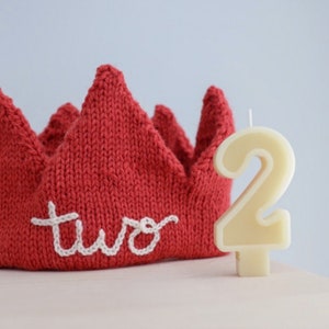 May include: A red knitted crown with the word "two" embroidered in white. Beside the crown is a cream-colored number "2" candle, perfect for a second birthday celebration.