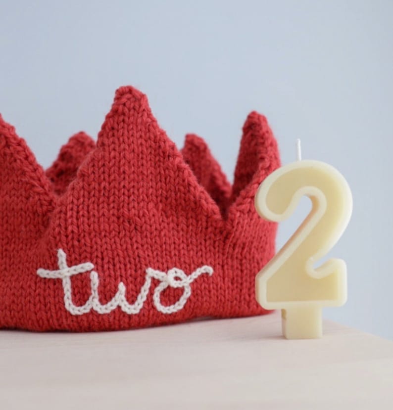 May include: A red knitted crown with the word "two" embroidered in white. Beside the crown is a cream-colored number "2" candle, perfect for a second birthday celebration.