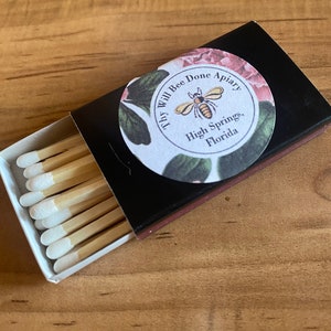 May include: A black matchbox with a floral label that reads "Thy Will Bee Done Apiary, High Springs, Florida." The box is open, revealing several wooden matches with white tips.