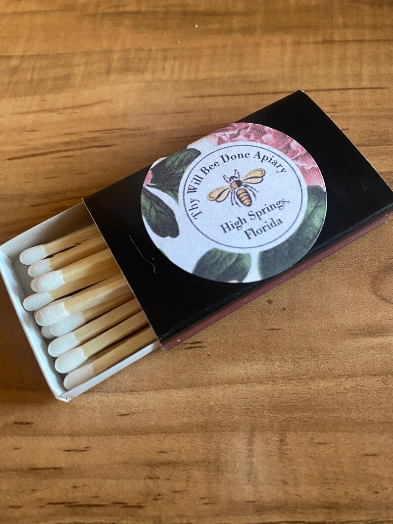 May include: A black matchbox with a floral label that reads "Thy Will Bee Done Apiary, High Springs, Florida." The box is open, revealing several wooden matches with white tips.