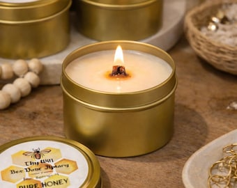 May include: A gold-coloured metal tin candle with a lit wooden wick. The label features a bee illustration, honeycomb patterns, and the text Thy Will Bee Done Jipmary Pure Honey 4oz Soy. Several identical closed candle tins are visible in the background.