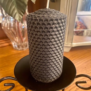 May include: A cylindrical charcoal gray candle featuring a textured honeycomb pattern across its entire surface. The candle has a single white cotton wick centered at the top. It is positioned upright on a black metal candle holder with curved legs.