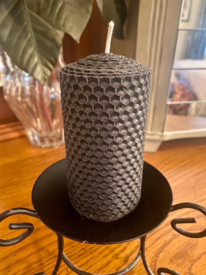 May include: A cylindrical charcoal gray candle featuring a textured honeycomb pattern across its entire surface. The candle has a single white cotton wick centered at the top. It is positioned upright on a black metal candle holder with curved legs.